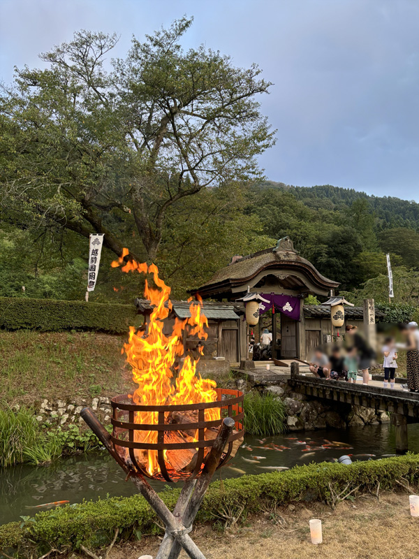 一乗谷朝倉館跡の唐門と篝火。手前に燃える篝火越しに、池に架かる橋と茅葺きの唐門が見える。