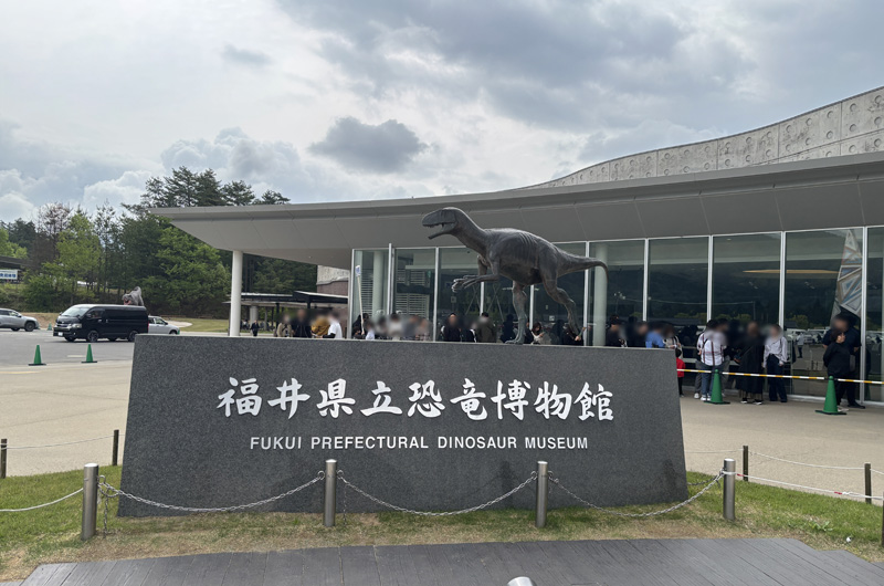 福井県立恐竜博物館の入口。恐竜のオブジェと特徴的なドーム型の建物、館名を刻んだ石碑が並ぶ