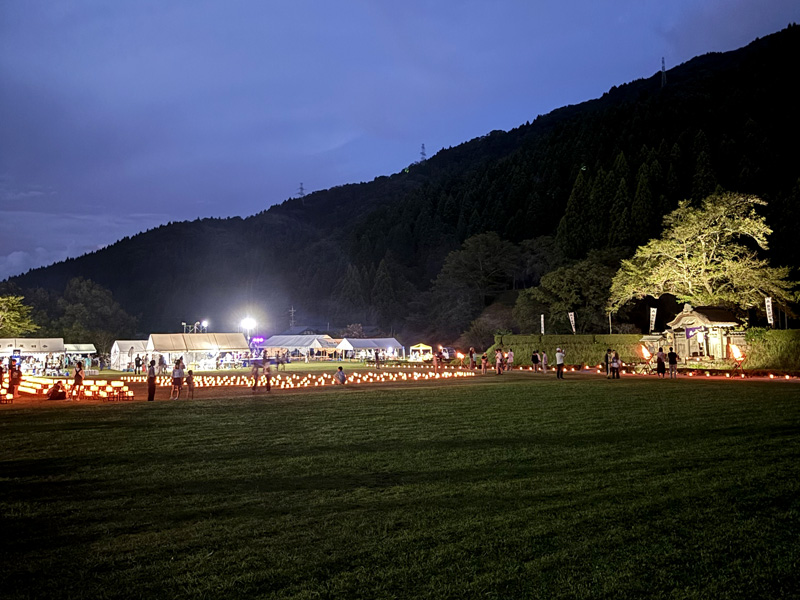 万灯夜の会場全景。夕暮れの空の下、キャンドルの灯りが会場一面に広がり、屋台やテントの明かりも見える。